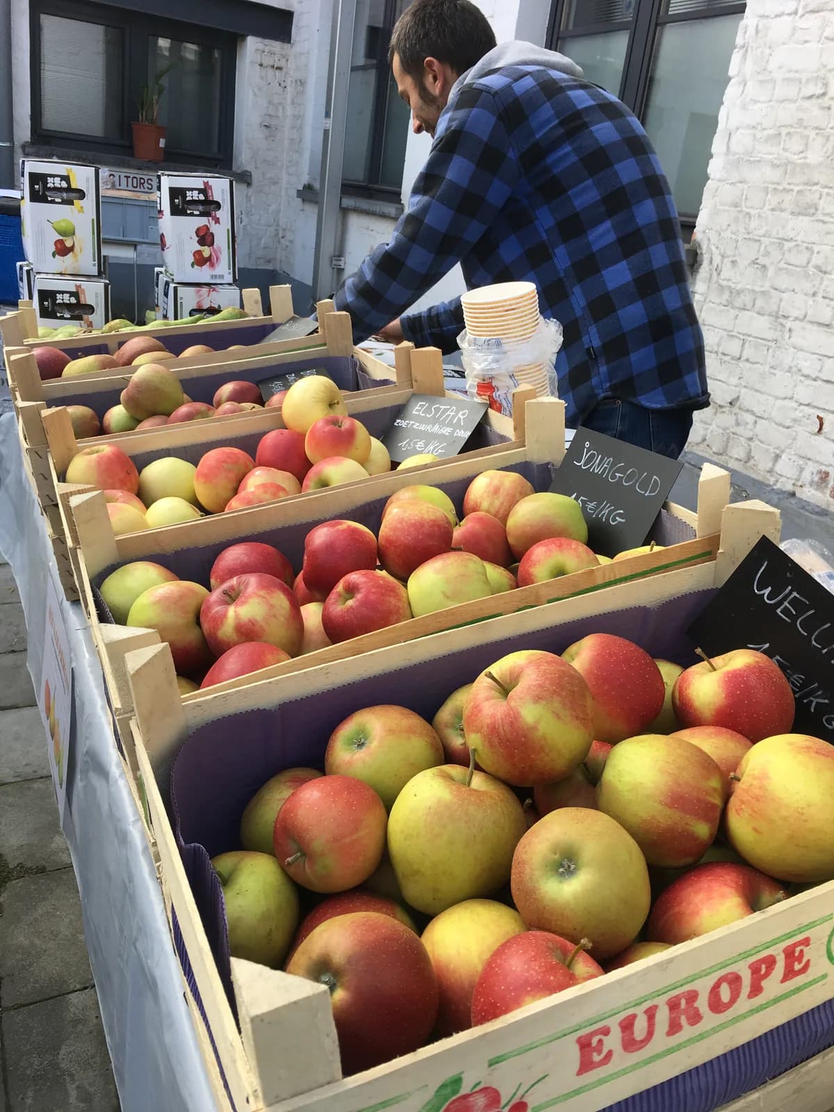 Des cagettes de pommes sur un étal de marché, devant des ardoises manuscrites.