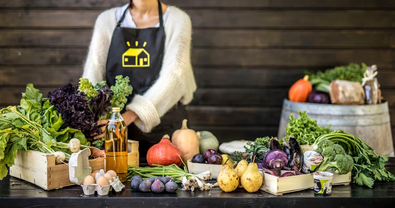 Une femme en tablier derrière un étal de légumes d'automne et de conserves.