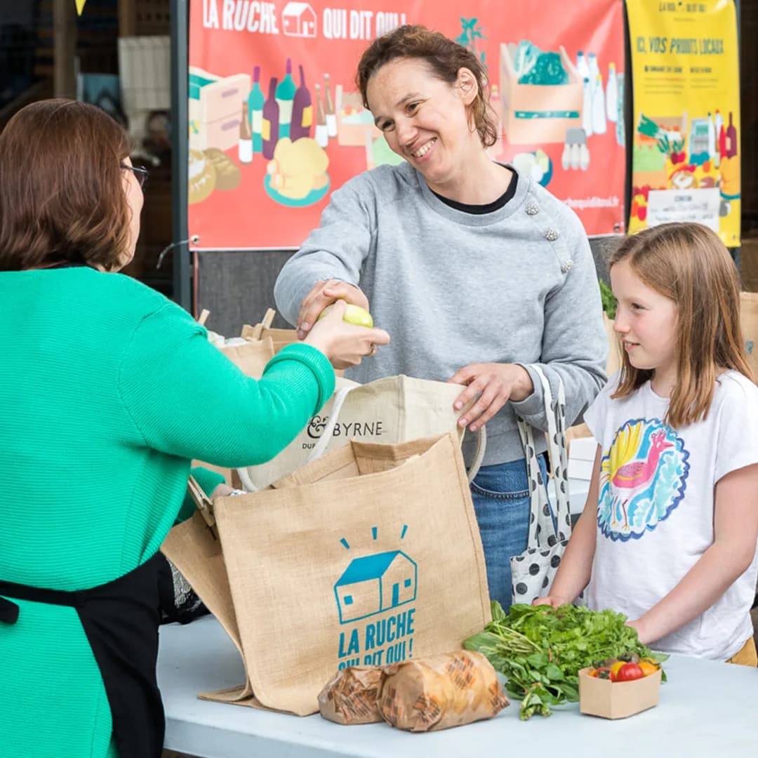 Une femme tend un sac en tissu rempli de légumes à une cliente, un enfant à ses côtés.