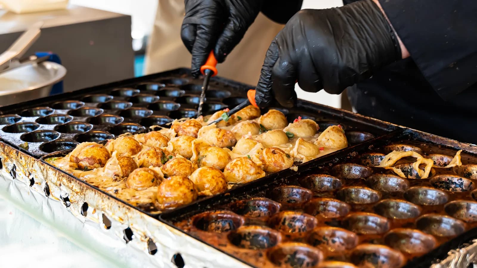Des takoyaki cuisent sur une plaque, retournés par des mains gantées.