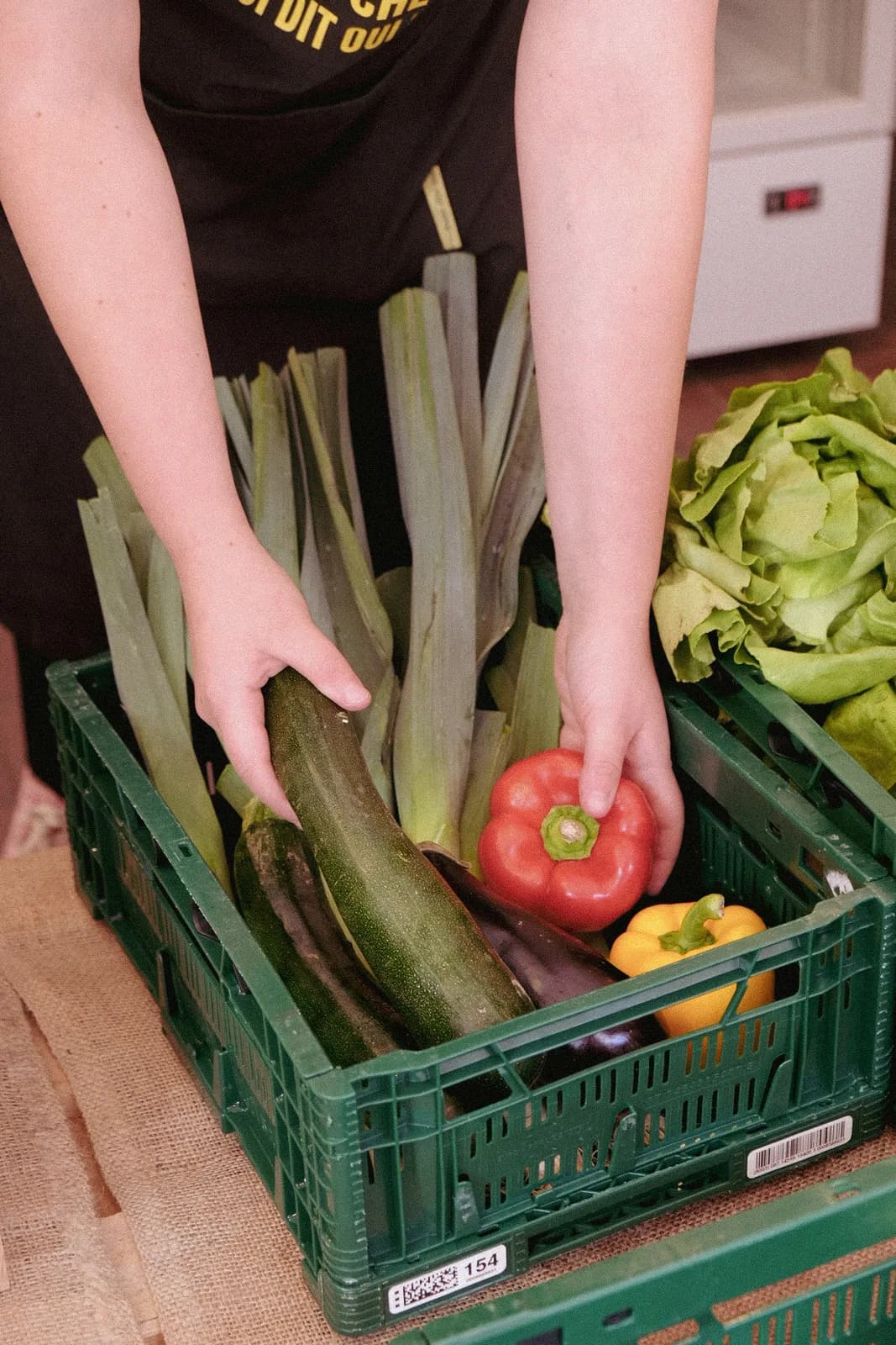 Des mains au-dessus de cagettes de légumes : courgettes, poivrons et salades.