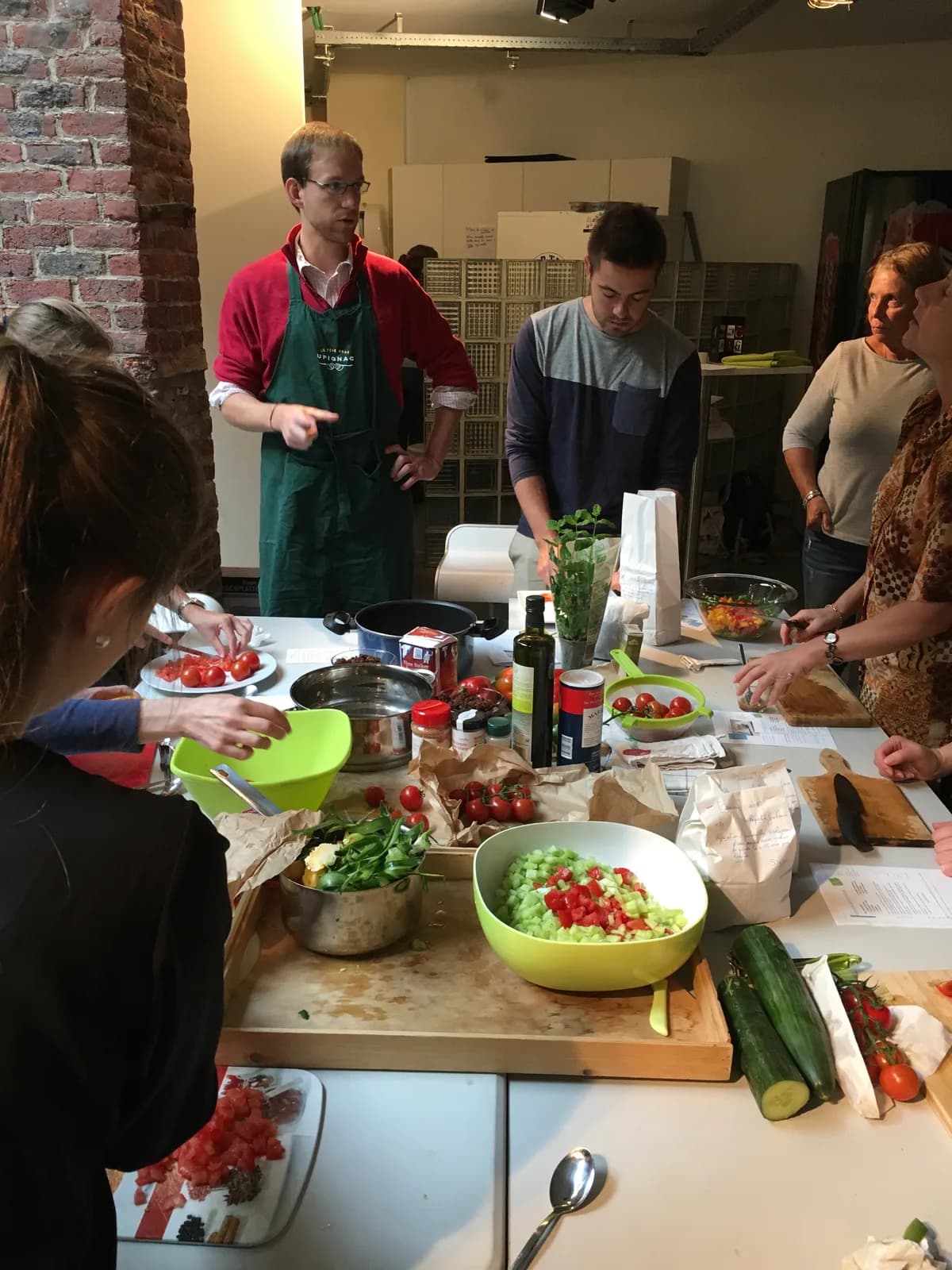 Un groupe prépare ensemble un repas autour d'une table : tomates, herbes, saladiers et planches à découper, en tablier.