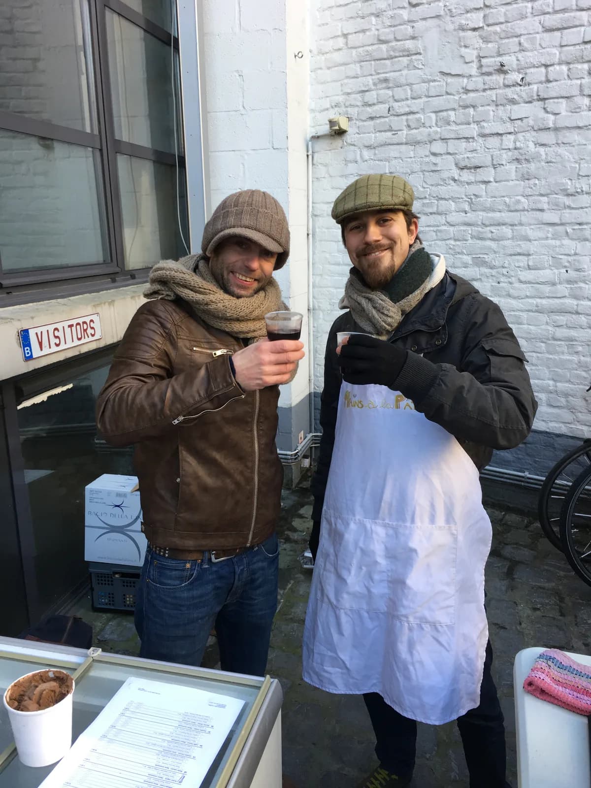 Deux membres de l'équipe en extérieur, l'un en tablier, une boisson chaude à la main.